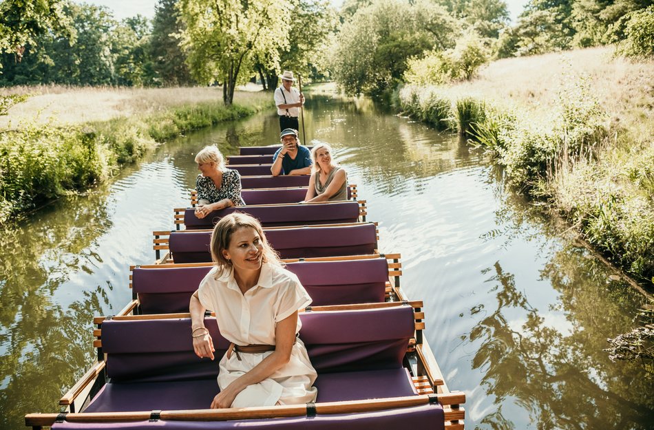 Frau sitz im Kahn vorne, hinten weitere Personen, werden durch den Spreewald gefahren.