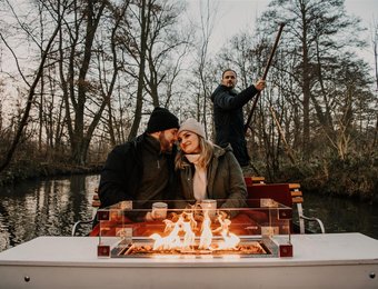 Ein Paar sitzt im Winter auf einem Kahn vor einem Kaminfeuer und wird durch den Spreewald gefahren.