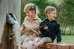 Zwei Kinder Mädchen und Junge sitzen auf dem Rand eines Brunnens und lachen während das Wasser hochspritzt