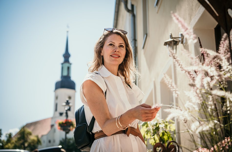 Frau steht vor Blumenladen und hält Blume in der Hand -  im Hintergrund eine Stadtszene mit Kirche