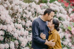 Mann und Frau umarmen sich und er küsst sie auf die Stirn im Hintergrund große rosa Blumensträucher