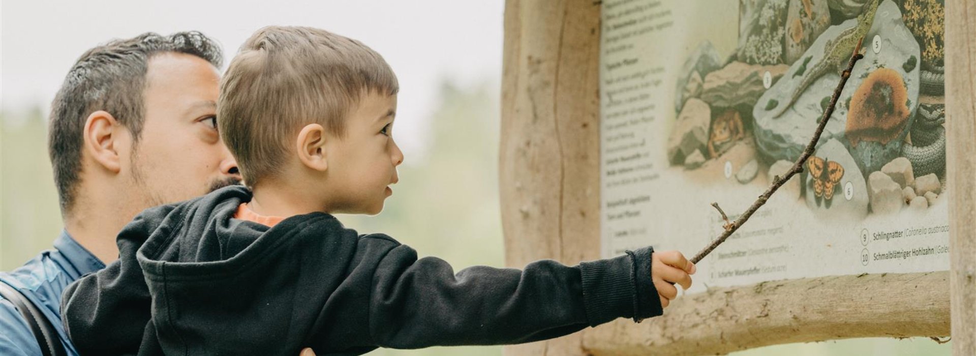 Mann hält Jungen hoch, der mit Stock auf Karte an Naturweg zeigt